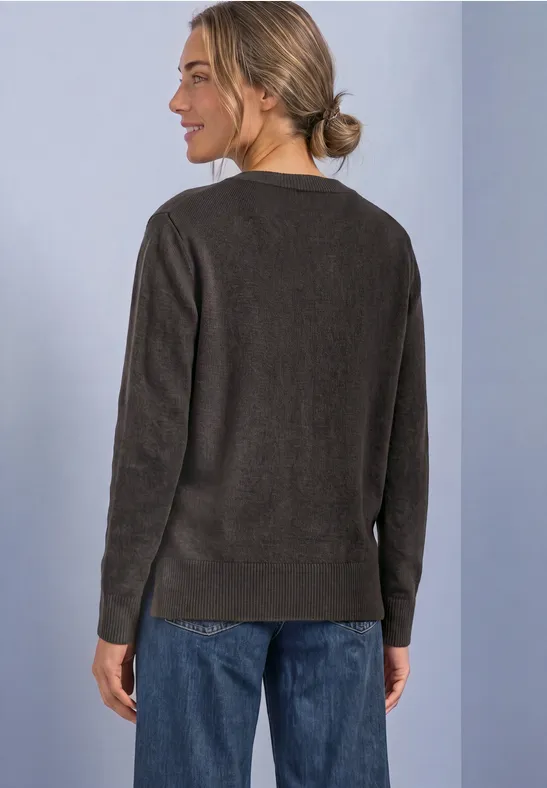 Femme vue de dos portant un pull marron foncé et un jean, cheveux en chignon, devant un mur clair.