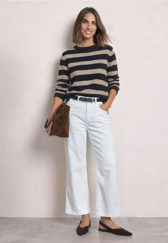 Frau in gestreiftem Pullover, weißen weiten Hosen und schwarzen Schuhen, hält braune Tasche.
