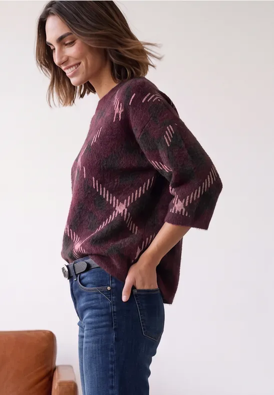 Frau in Burgunderfarbenem Pullover mit Rautenmuster, Jeans und Gürtel, lächelnd seitlich posiert.