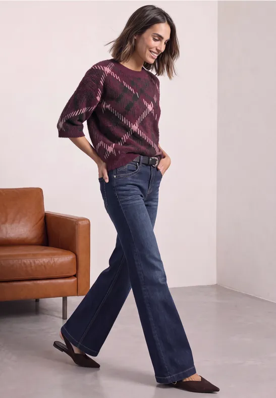 Frau in karierter Bordeaux-Pulli und ausgestellten Jeans posiert neben braunem Ledersofa.