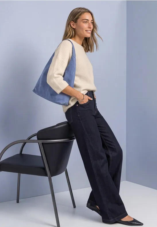 Frau mit weitem Jeans, hellblau-beige Oberteil und blauer Tasche lehnt an Stuhl vor minimalistischem Hintergrund.