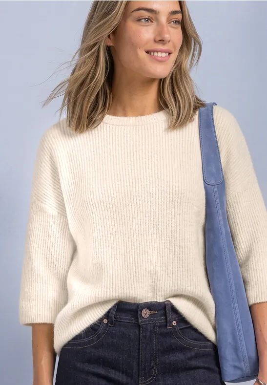 Frau lächelt in hellem Pullover mit 3/4-Ärmeln, dunkle Jeans und blaue Schultertasche vor hellem Hintergrund.