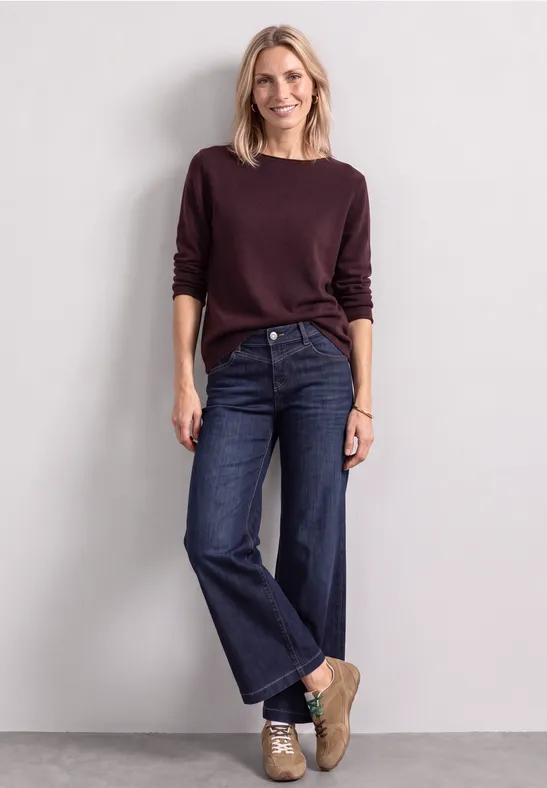 Eine Frau mit blondem Haar trägt Burgunder-Pullover, weite Jeans und Sneaker vor neutralem Hintergrund.