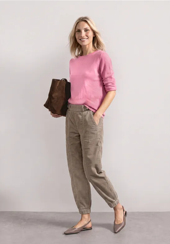 Vrouw in roze trui, kaki cargobroek en bruine clutch staat voor een lichte achtergrond.