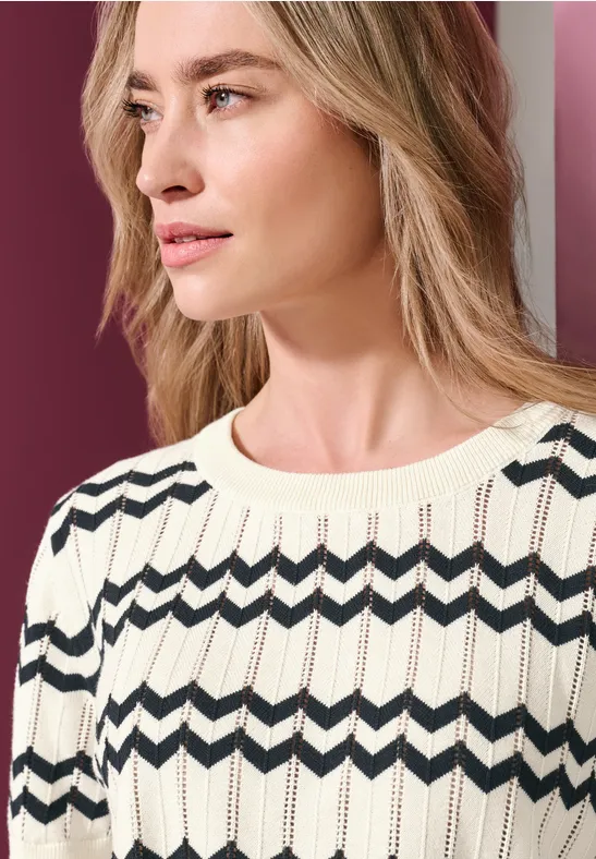 Frau in hellem gestrickten Pullover mit dunklem Zickzackmuster, Blick seitlich nach rechts.