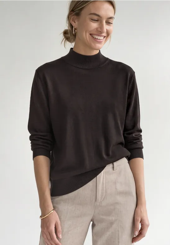 Une femme souriante en pull noir à col roulé et pantalon clair sur fond blanc.