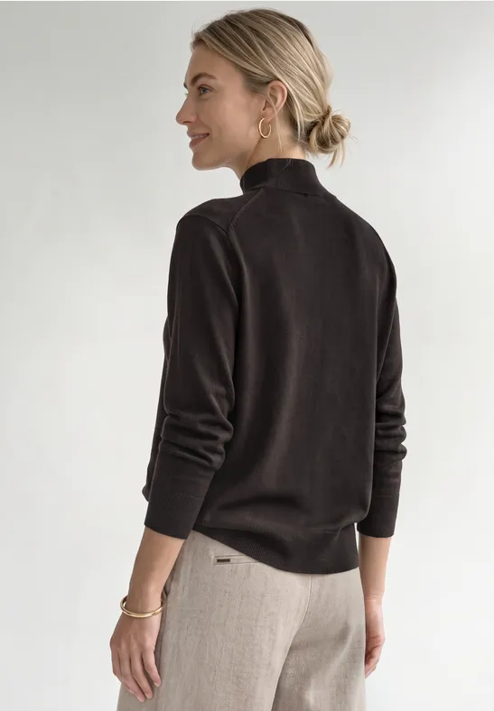 Une femme vue de dos porte un pull col roulé sombre et un pantalon clair, cheveux relevés, fond épuré.