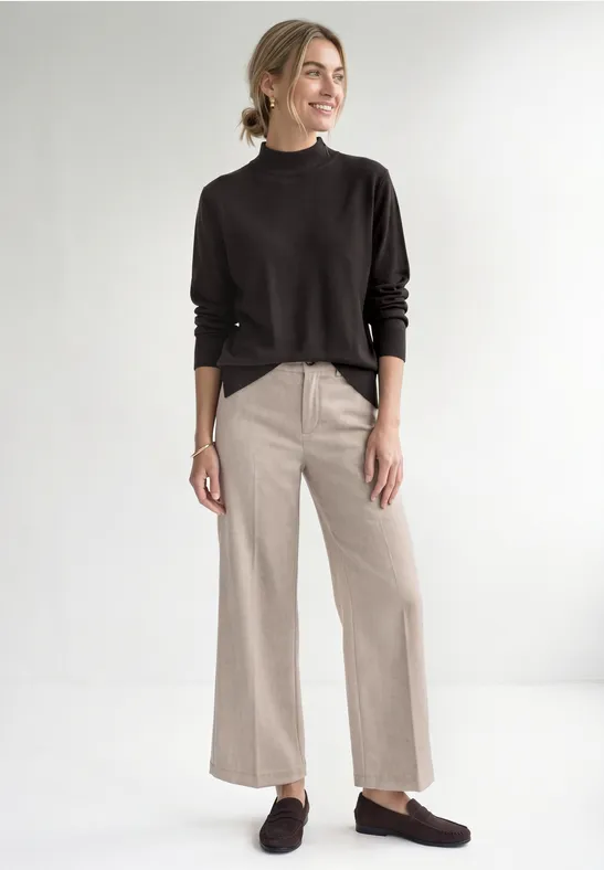 Femme devant un fond blanc, porte un pull noir, un pantalon large beige et des mocassins marron.