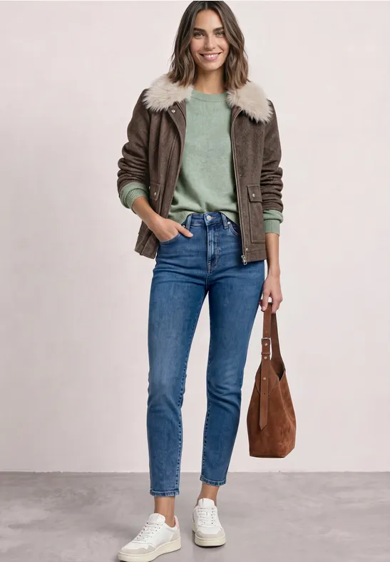 Junge Frau in blauen Jeans, grünem Pullover, brauner Lammfelljacke, weißen Sneakern und brauner Tasche.