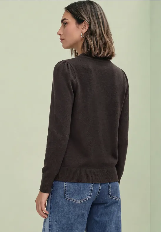 Frau im Profil trägt braunen Pullover und Jeans vor schlichtem, hellem Hintergrund.