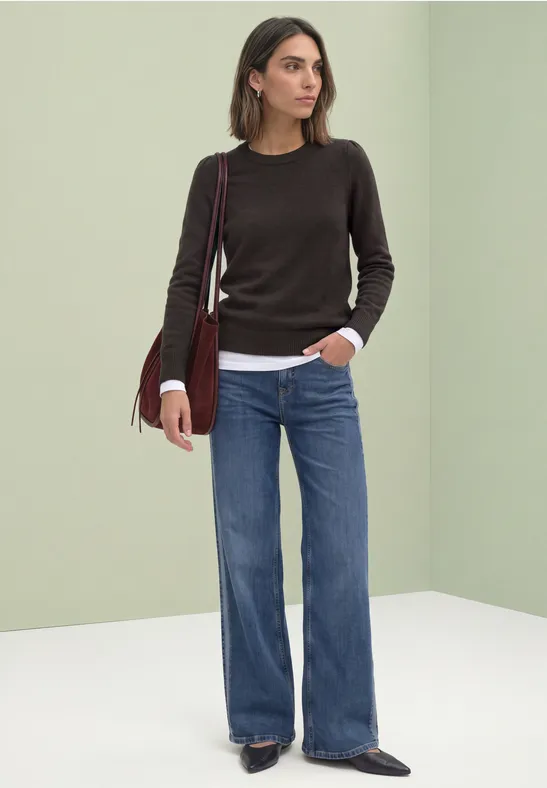 Eine Frau trägt einen dunklen Pullover, weiße Bluse, weite Jeans und rote Umhängetasche vor hellem Hintergrund.