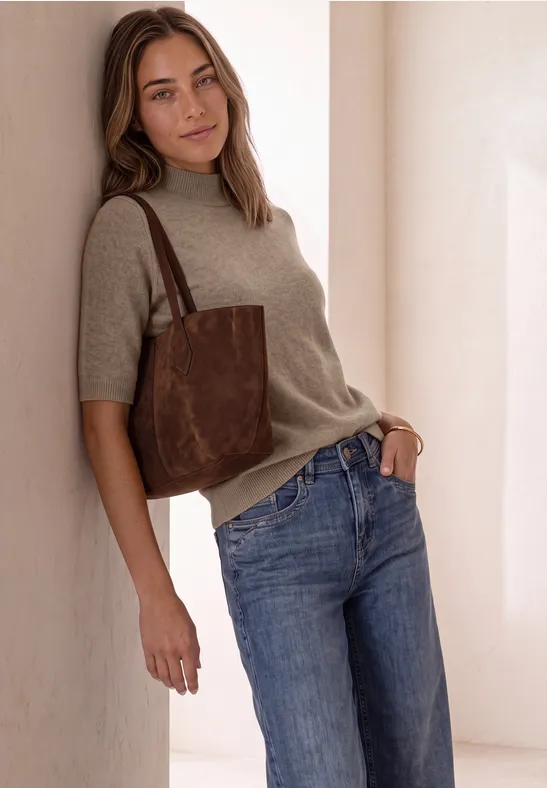 Junge Frau lehnt an heller Wand, trägt beigefarbenen Pullover, blaue Jeans und braune Schultertasche.