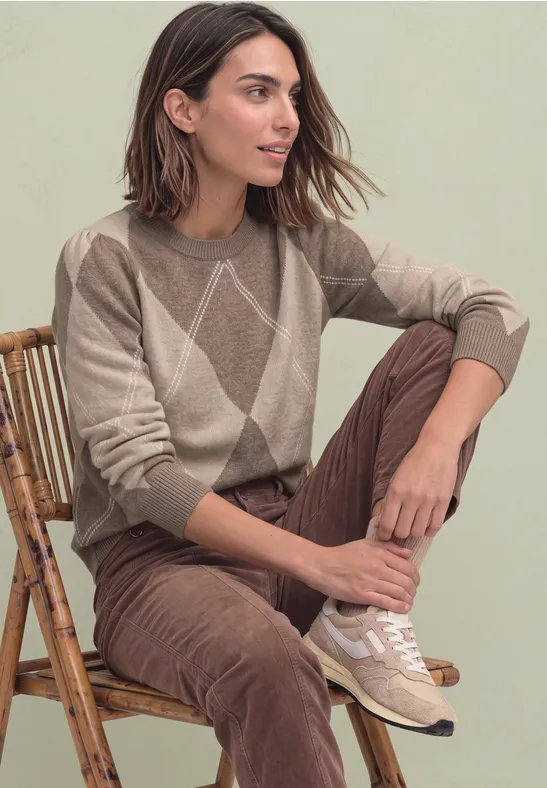 Eine Frau sitzt auf einem Rattanstuhl, trägt einen beigen Pullover mit Rautenmuster und braune Cordhose.