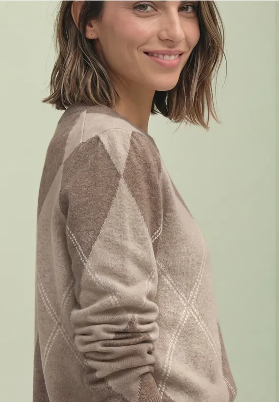 Eine lächelnde Frau mit schulterlangen braunen Haaren trägt einen beigen Rautenmuster-Pullover vor hellem Hintergrund.