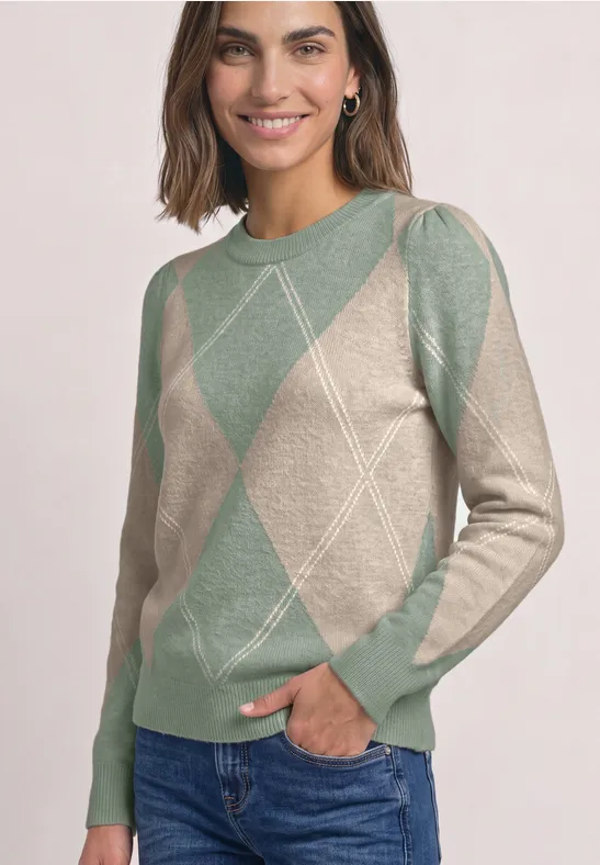 Vrouw glimlacht in beige-groene ruitjestrui met lange mouwen en jeans voor lichte achtergrond.