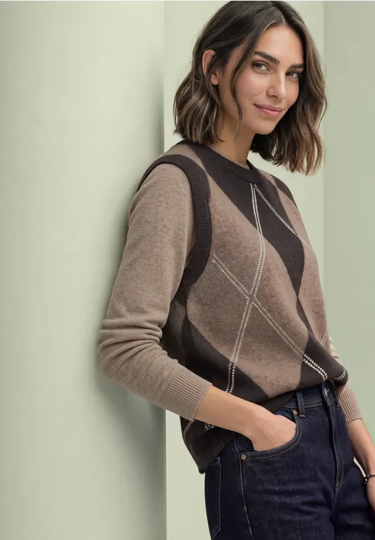 Eine Frau lehnt lässig an einer Wand, trägt einen braunen Strickpullover mit Rautenmuster und dunkle Jeans.