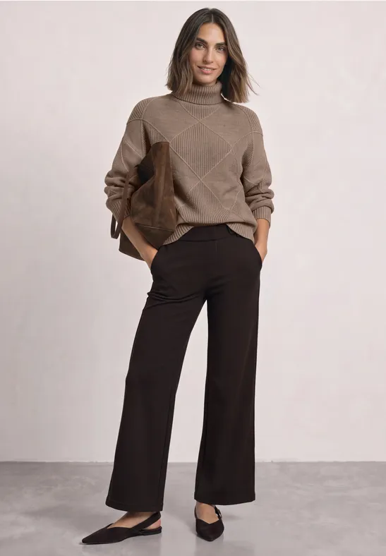 Frau trägt braunen Pullover und weite dunkelbraune Hosen, schwarze Ballerinas und hält braune Tasche.
