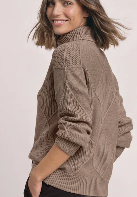 Eine lächelnde Frau trägt einen beigen, strukturierten Strickpullover mit Stehkragen und blickt über die Schulter.