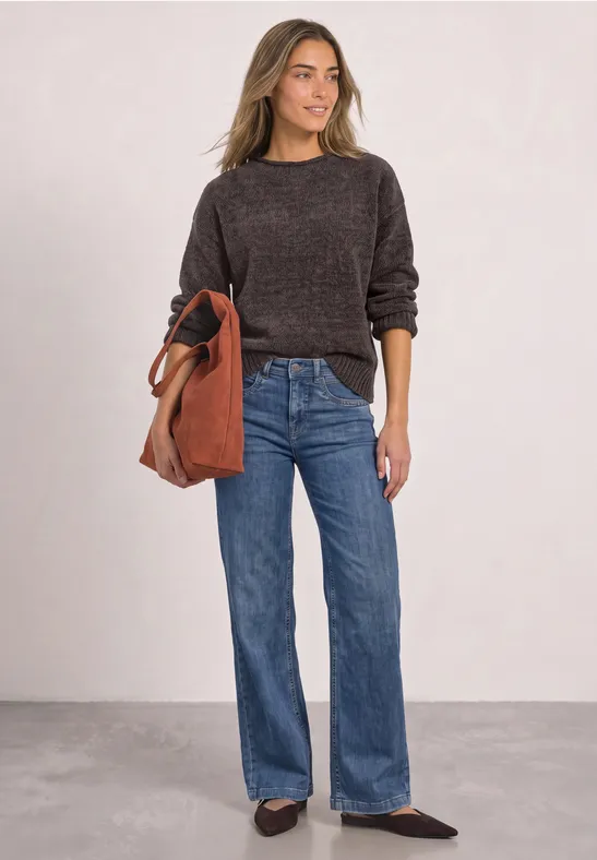 Frau im braunen Pullover, weitem Jeans, Slingback-Schuhen und roter Ledertasche vor neutralem Hintergrund.