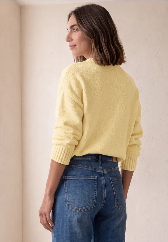Femme de dos en pull jaune et jean, debout de profil sur fond neutre.