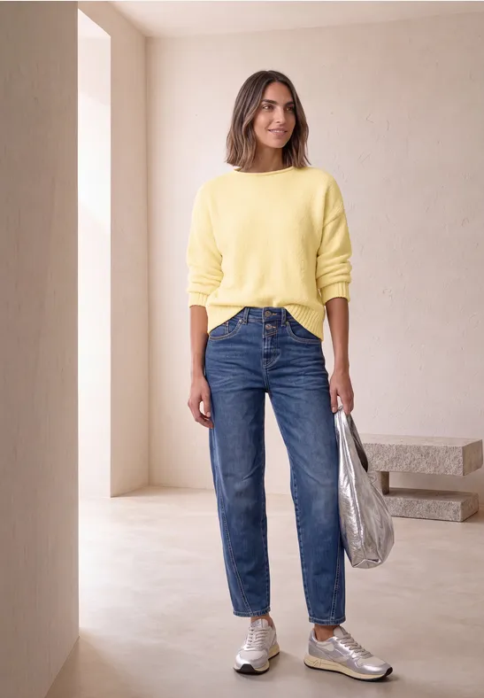 Femme dans une pièce minimaliste portant un pull jaune, un jean bleu, des baskets argentées et un sac argenté.
