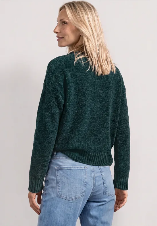 Femme blonde vue de dos, portant un pull vert foncé et un jean, regard de profil.