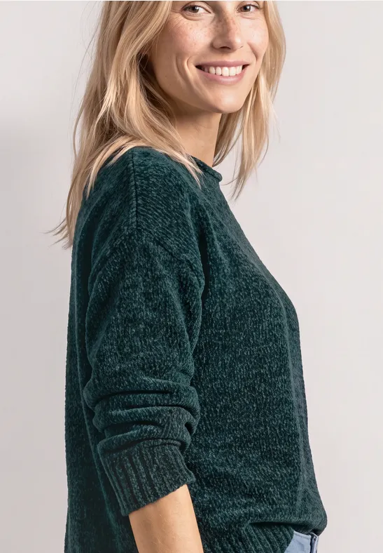 Portrait d’une femme souriante aux cheveux clairs portant un pull vert foncé moelleux sur fond neutre.