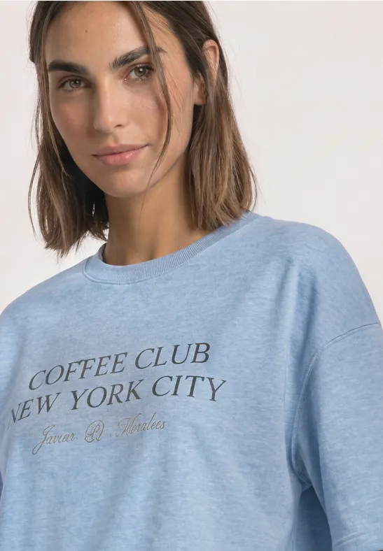 Kobieta w jasnoniebieskiej bluzie z napisem "Coffee Club New York City", lekki uśmiech