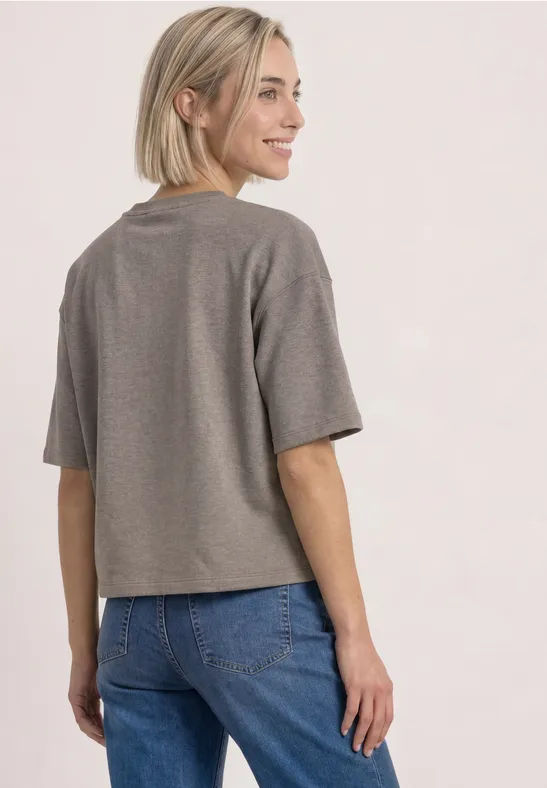 Frau schaut lächelnd über die Schulter, trägt graues, kurzes T-Shirt und blaue Jeans vor neutralem Hintergrund.