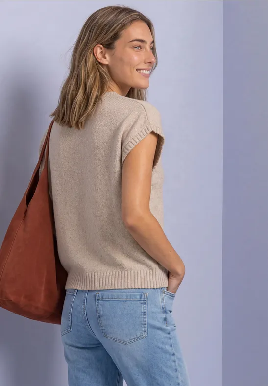 Junge Frau in beigem Stricktop, Jeans und brauner Tasche schaut lächelnd über die Schulter.