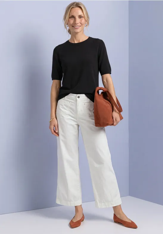 Frau in schwarzem T-Shirt, weißen weiten Jeans und braunen Ballerinas mit großem braunen Beutel vor lila Wand.