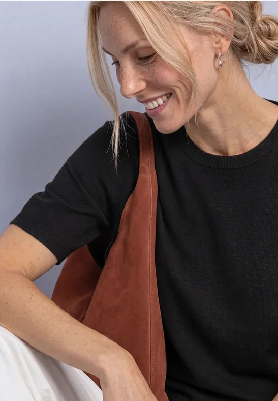 Junge Frau mit blondem Haar trägt schwarzes T-Shirt und braune Schultertasche, lächelt seitlich.