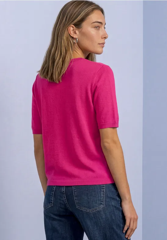Frau von hinten in pinkem Kurzarm-Pullover und Jeans vor hellem Hintergrund.