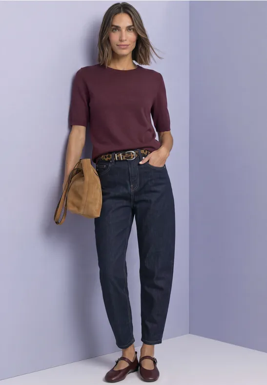 Femme en pull violet à manches courtes, jean foncé, ballerines marron et sac beige devant un mur lilas.