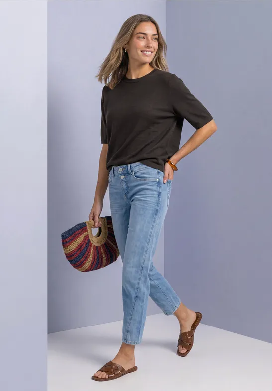 Frau in schwarzem T-Shirt, hellblauen Jeans und braunen Sandalen lächelnd mit bunter Tasche.