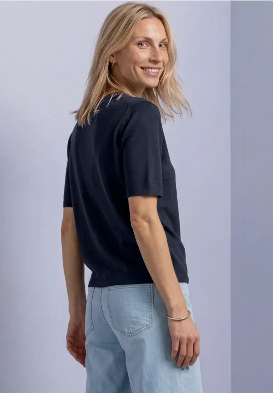 Blonde Frau in schwarzem Shirt und Jeans, die lächelnd über die Schulter blickt.