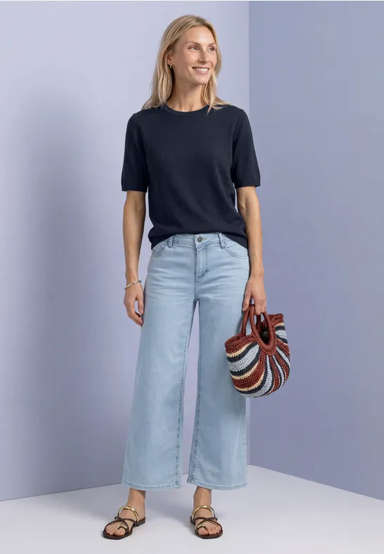 Frau in schwarzem T-Shirt, weiten, hellen Jeans und Sandalen hält gestreifte Handtasche.