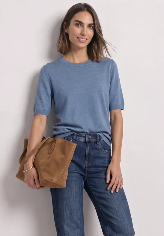 Een vrouw draagt een blauw shirt met korte mouwen, een spijkerbroek en houdt een bruine tas vast.