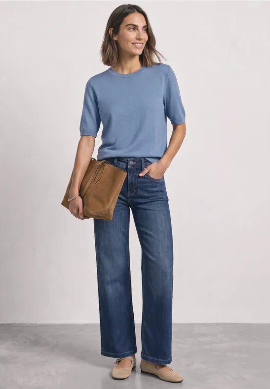 Een vrouw draagt een blauw T-shirt, wijde jeans en houdt nonchalant een bruine tas onder haar arm.