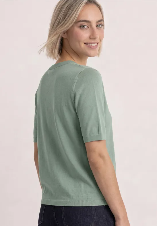 Frau im hellgrünen T-Shirt zeigt Blick über die Schulter und lächelt.