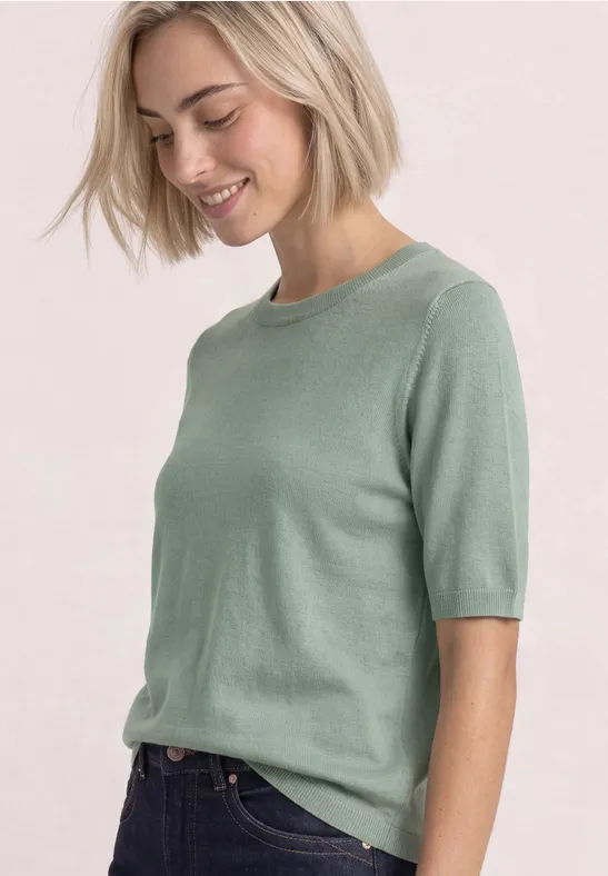 Lächelnde Frau mit kurzen blonden Haaren, grünes Kurzarmshirt und Jeans.