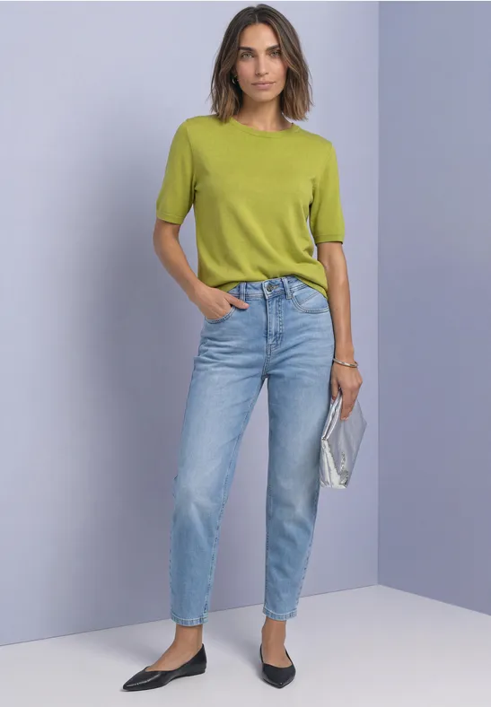 Frau in grünem Kurzarmshirt und blauen Jeans, hält silberne Clutch und trägt schwarze Ballerinas.