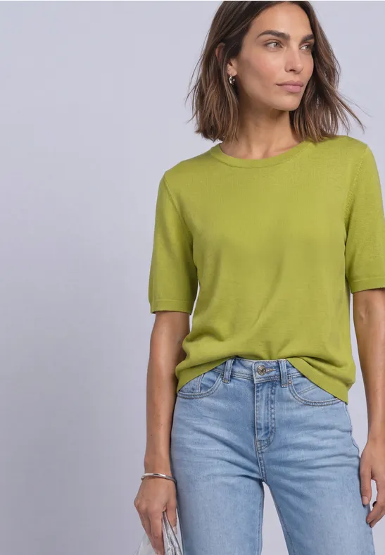 Eine Frau mit schulterlangem braunem Haar trägt ein hellgrünes T-Shirt und blaue Jeans vor hellem Hintergrund.
