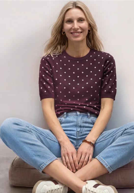 Blonde Frau im Schneidersitz vor weißer Wand, trägt gepunktetes lila Shirt und blaue Jeans.