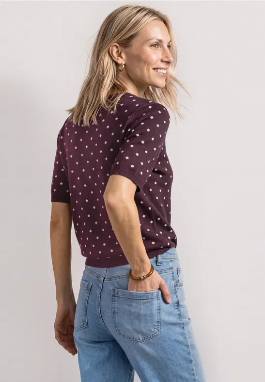 Eine Frau in blauer Jeans und gepunktetem T-Shirt blickt lächelnd seitlich über die Schulter.