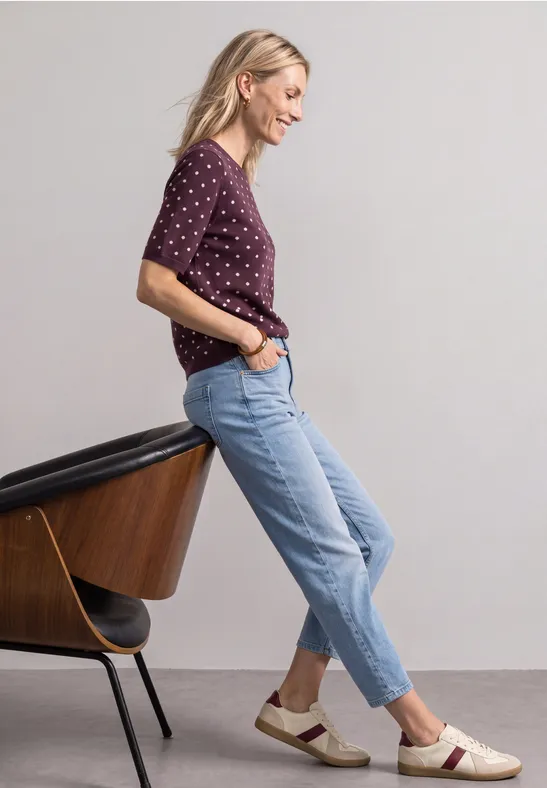 Frau lehnt seitlich an Stuhl, trägt lila gepunktetes Shirt, helle Jeans und bequeme Loafers.