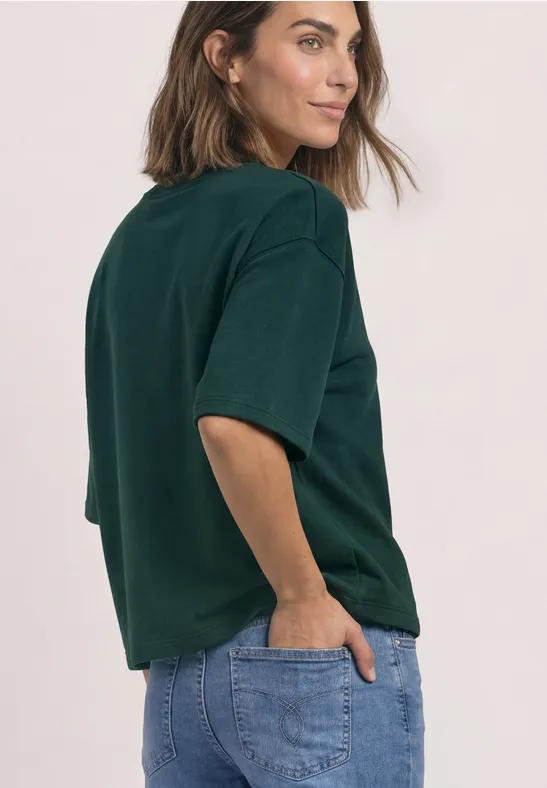 Kobieta z boku w zielonym t-shircie i niebieskich dżinsach, ręce w kieszeniach.