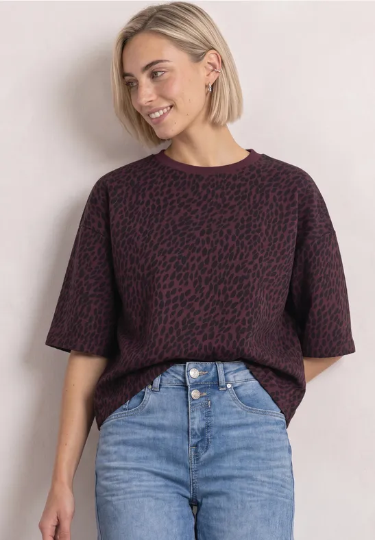 Jeune femme au carré blond porte un pull court violet à motifs et un jean taille haute, devant un mur clair.