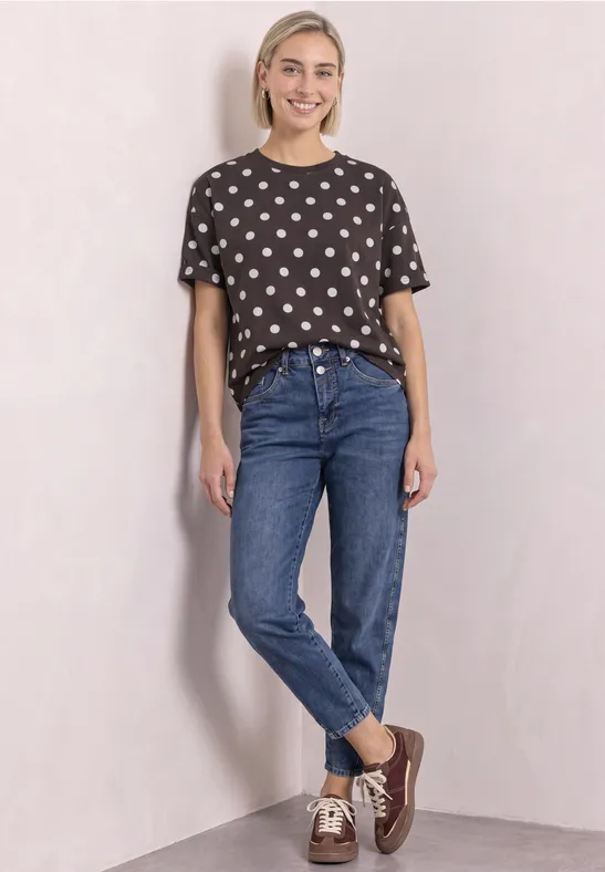 Junge Frau in gepunktetem T-Shirt und Jeans steht lächelnd an heller Wand.
