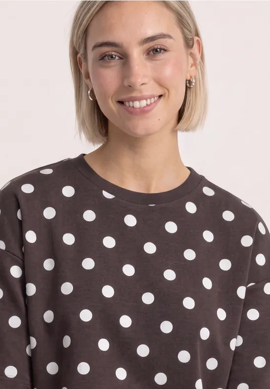 Junge Frau mit kurzem blondem Haar lächelt und trägt einen braunen Pullover mit weißen Punkten.